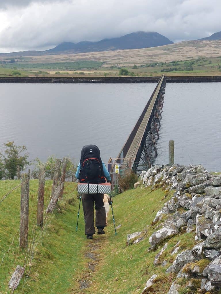 Rear view of a backpacker with a large black pack walking down a grassy path toward a long, narrow footbridge spanning a wide lake in Wales.