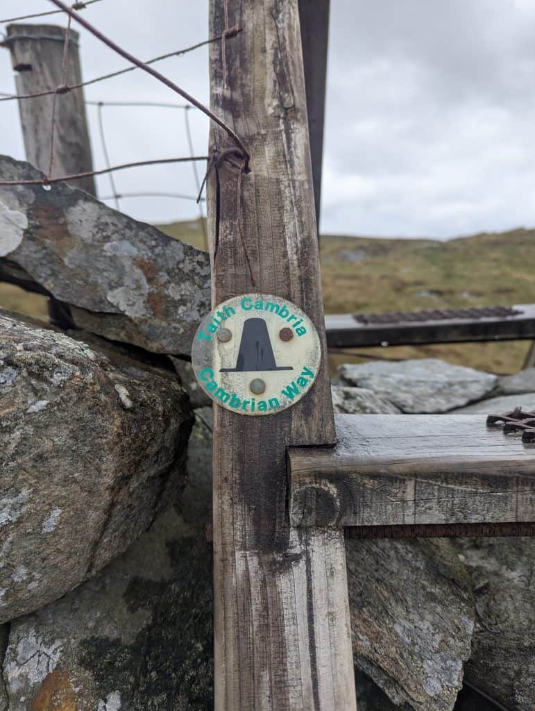 Close up of a circular Cambrian Way trail marker disc attached to a wooden ladder stile next to a stone wall in Eryri National Park, Wales.