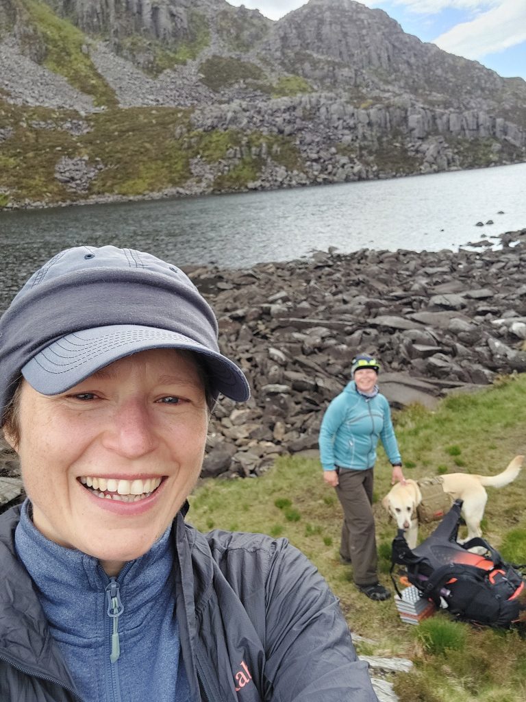 Close-up selfie of a smiling hiker with a companion and yellow dog Poppy standing by a boulder-lined mountain lake in the Rhinogydd range, Wales.