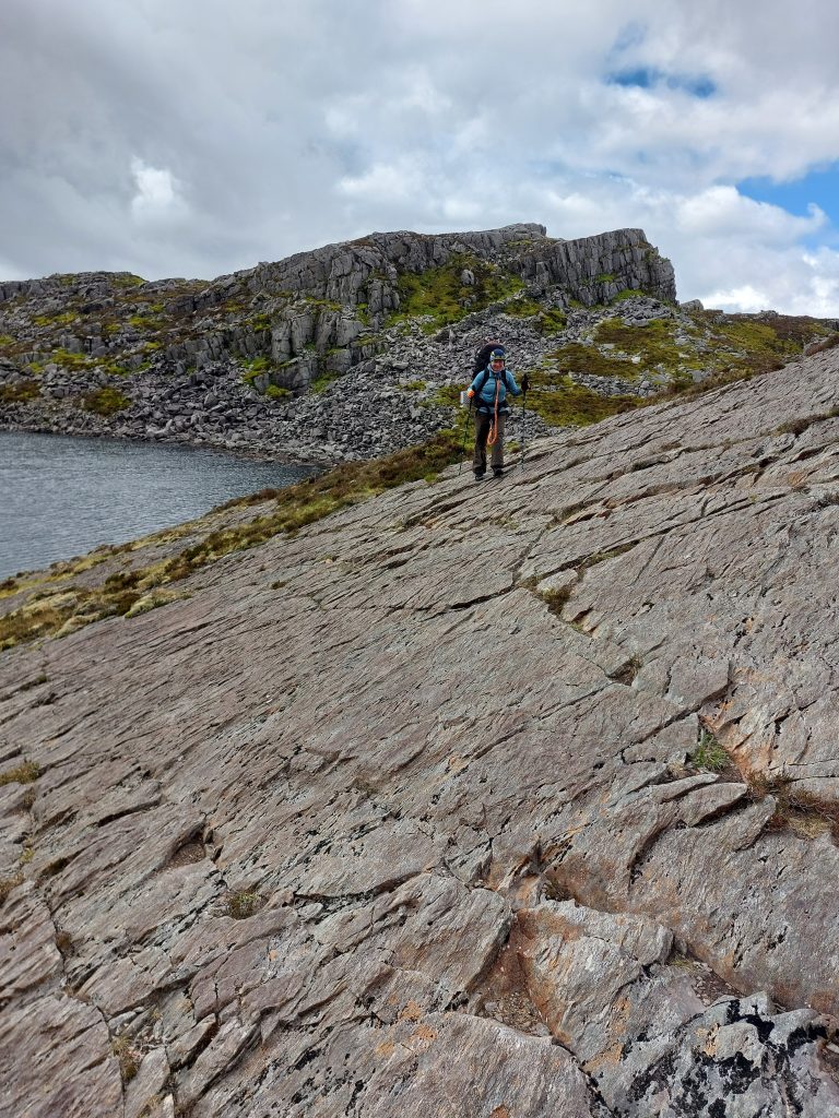 Hiker using trekking poles walks across a wide, sloping rock slab alongside a mountain tarn on the Rhinogydd Traverse path in Wales.