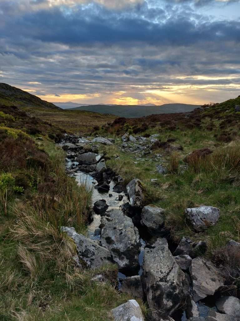 A rocky mountain stream cuts through the grassy heather moorland of the Rhinogydd range in Wales as sunset rays break through dramatic dark clouds over distant hills.