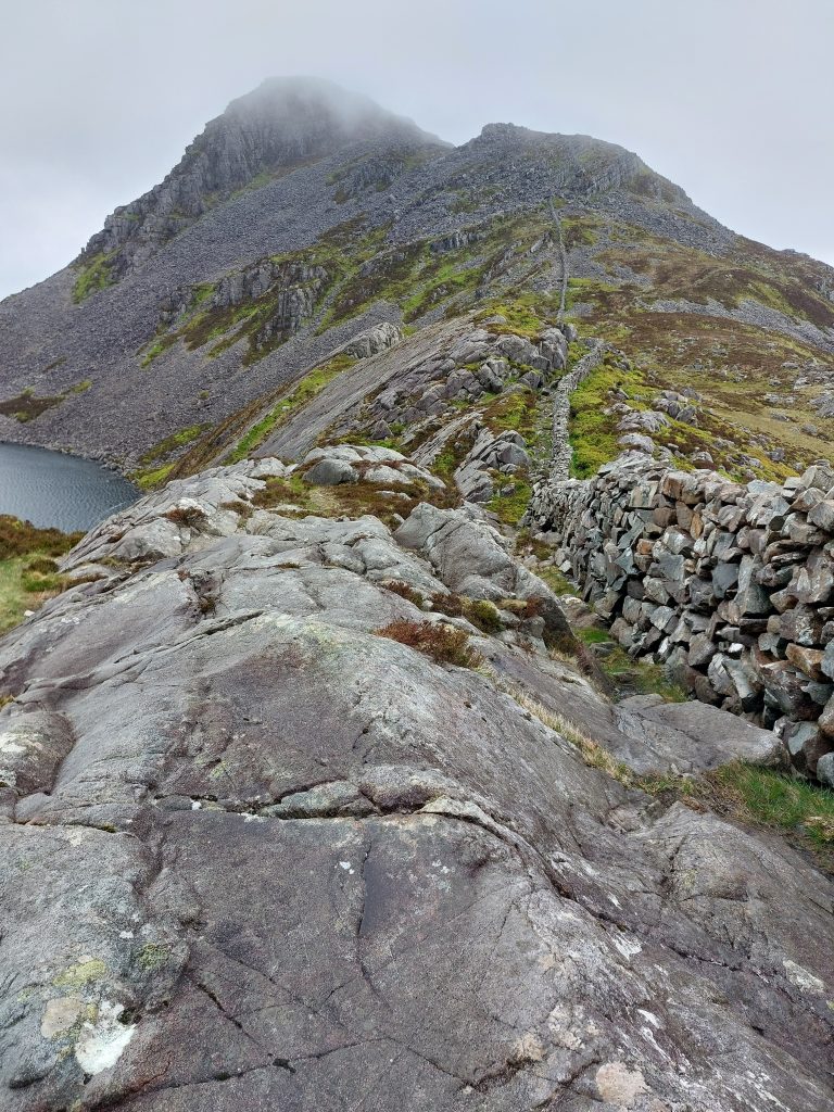 A steep rocky mountain ridge with a dry stone wall climbing the technical boulder slope toward a misty peak next to a deep tarn in Wales.