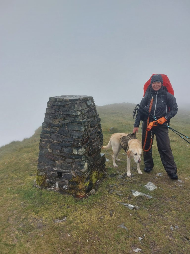 Hiker in wet weather gear and yellow dog Poppy standing next to a stone trig pillar on a foggy mountain peak with zero visibility.