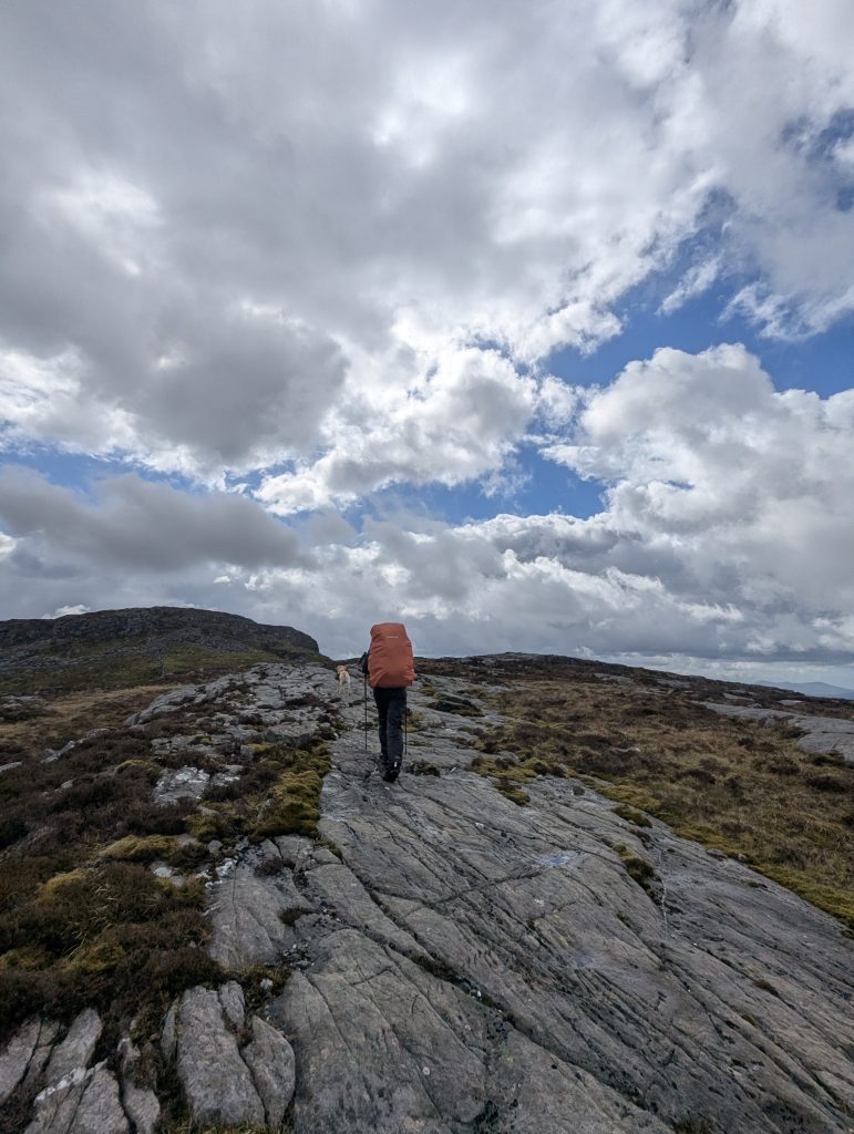 A backpacker with a large orange pack cover hikes up a broad, glaciated rock slab on a wild mountain ridge with trail dog Poppy under a dramatic cloudy sky in Wales.