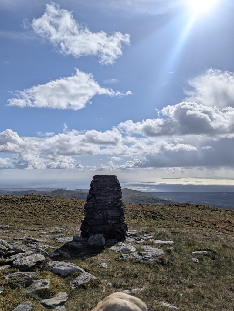 A traditional stone-built mountain trig pillar stands on a grassy summit overlooking a vast, sunlit coastline and the sea under a bright blue sky with dramatic white clouds.