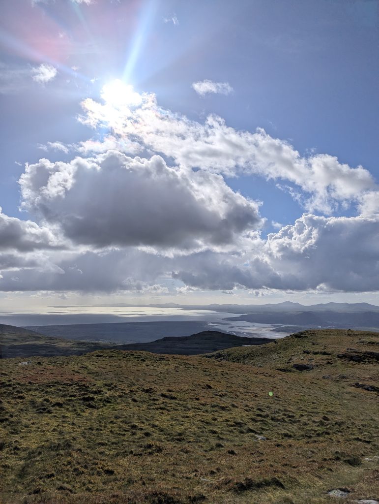 Wide landscape view from a grassy mountain slope looking down onto a sweeping coastal river estuary and the sea as bright sunbeams burst through dramatic white clouds in Wales.