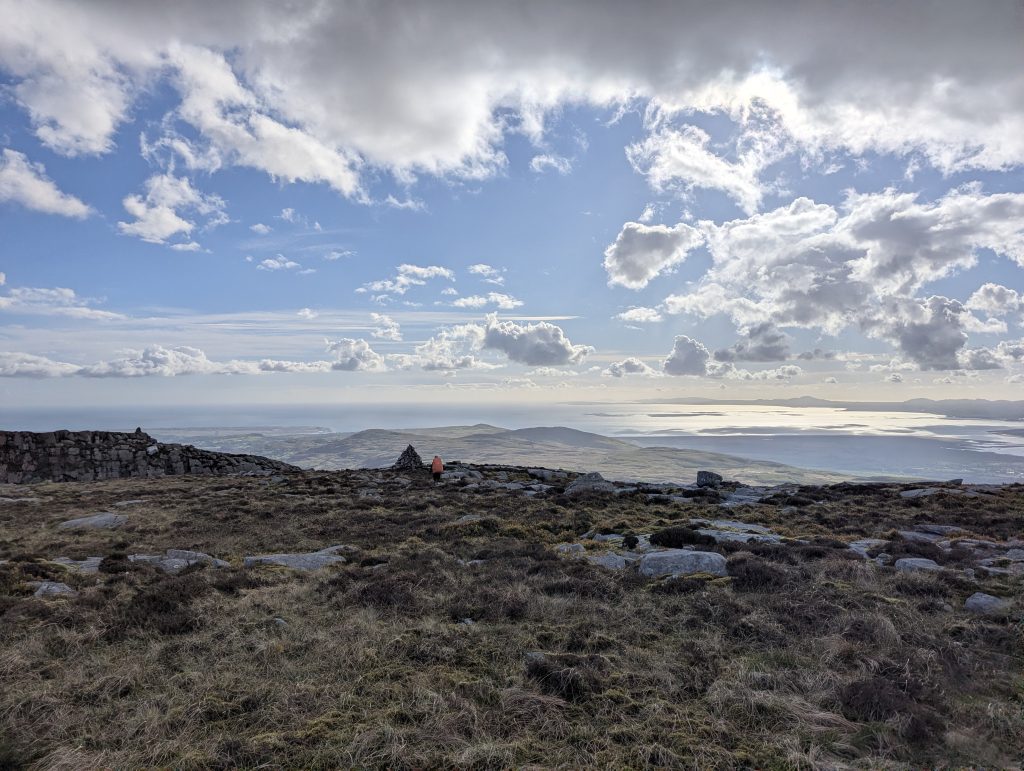 A distant backpacker with an orange pack cover walks past a small stone cairn on a vast, grassy mountain ridge overlooking a sunlit coastal estuary in Wales.