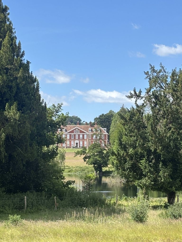 A grand, historic English manor house partially hidden behind lush green trees along the trail.