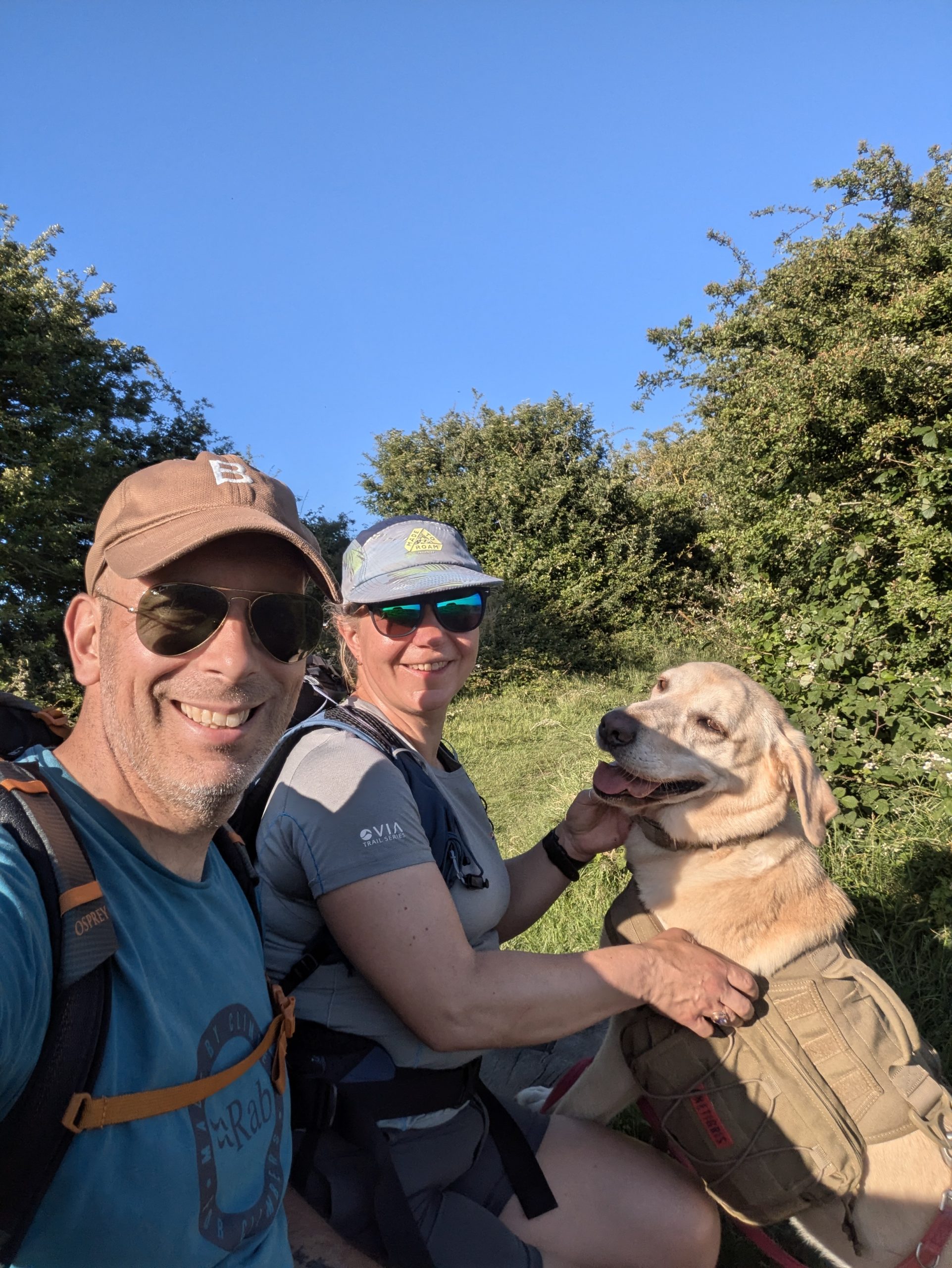 A close-up selfie of two smiling hikers wearing sunglasses, caps, and trekking backpacks alongside a yellow Labrador cross wearing a tan dog harness, resting on a sunny, green trail bordered by hedgerows on the Elham Valley Way.