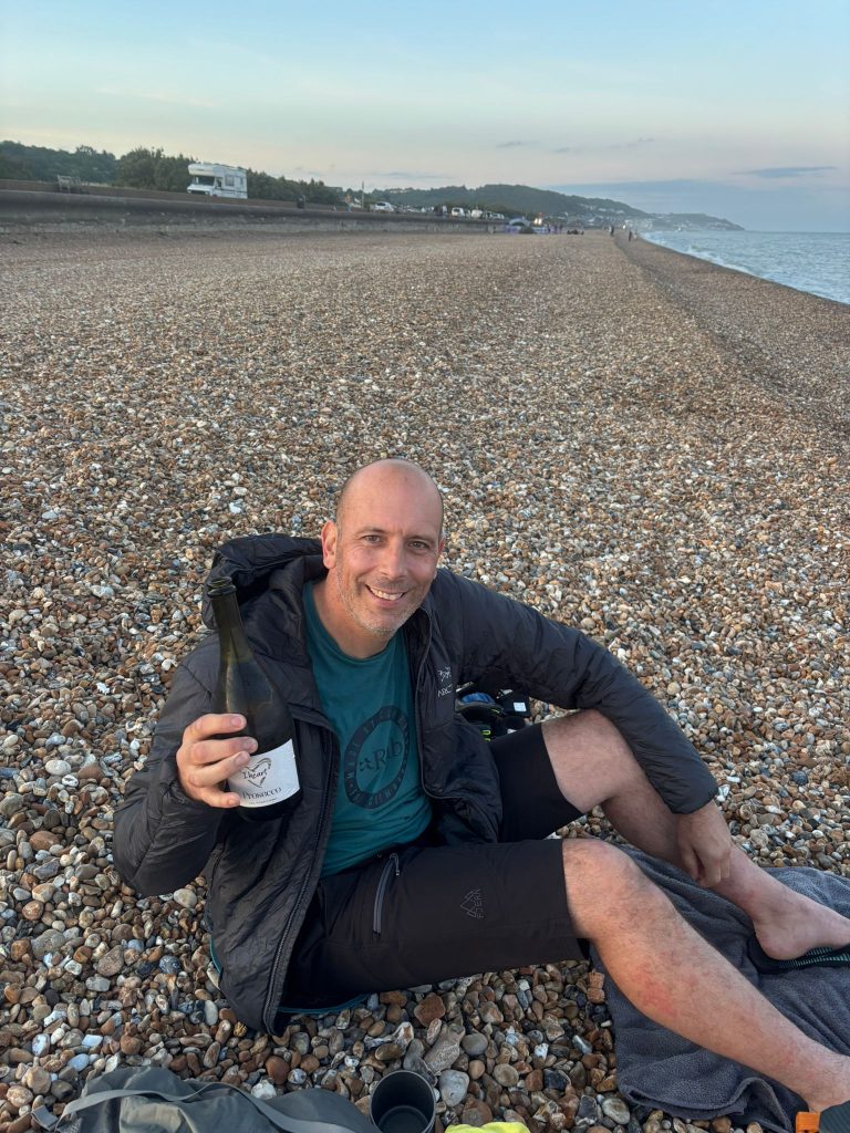 A hiker sitting on a pebble beach smiling while opening a bottle of prosecco.