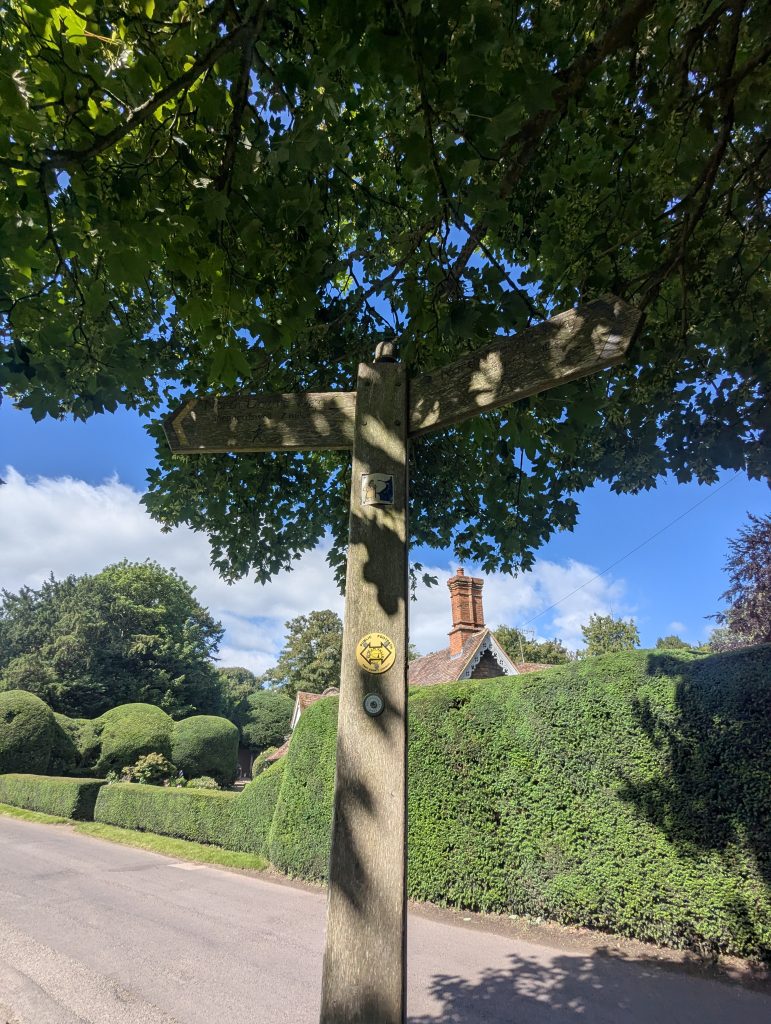 A green public footpath sign pointing off the paved road and into the Kent countryside.