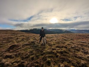 Hiker Syla stands alongside her yellow Labrador cross Poppy on the wide, grassy summit ridge of Broom Fell in the Lake District, silhouetted against a dramatic winter sky with a bright low sun bursting through rolling mountain clouds.