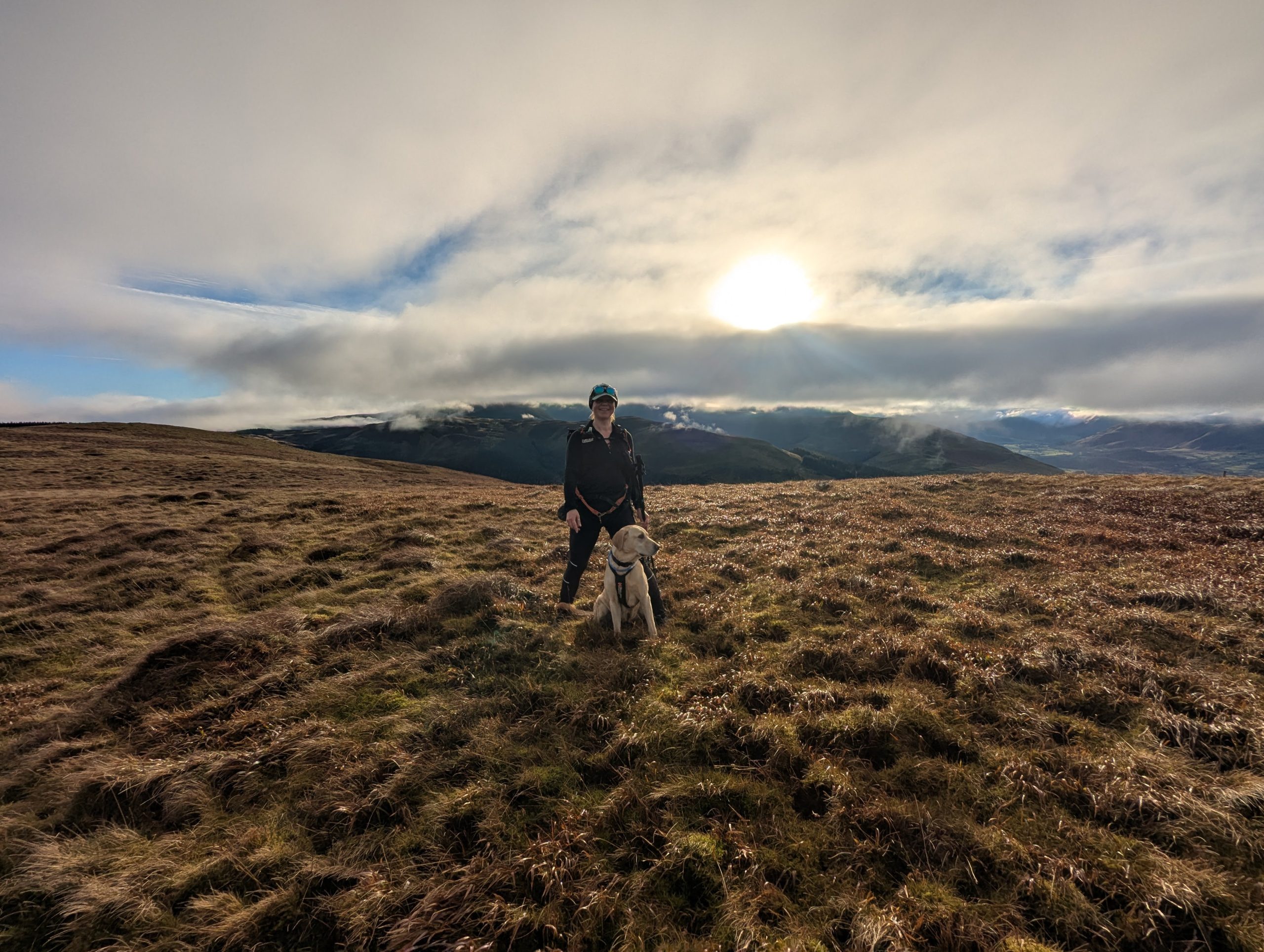 Hiker Syla stands alongside her yellow Labrador cross Poppy on the wide, grassy summit ridge of Broom Fell in the Lake District, silhouetted against a dramatic winter sky with a bright low sun bursting through rolling mountain clouds.