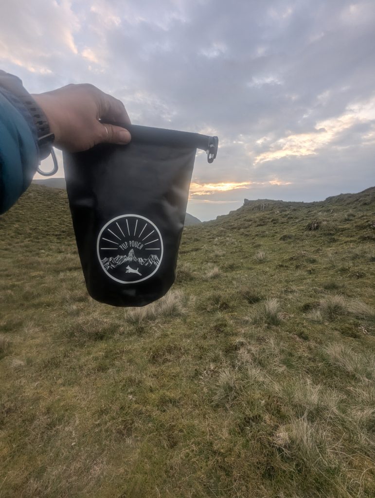 A hand holding a black waterproof roll-top dog waste storage bag featuring a white circular emblem of a bounding dog over mountains, silhouetted against a golden upland sunset valley.