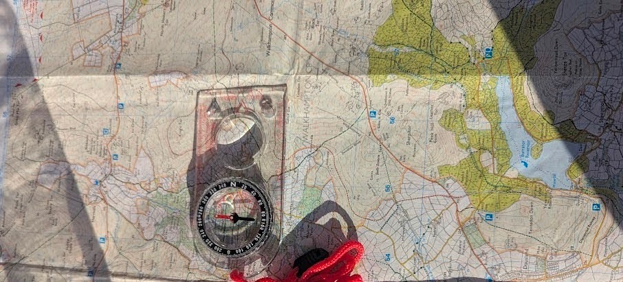Close-up of a physical UK Ordnance Survey Explorer map unfolded on a wooden surface with a clear plastic baseplate compass aligned over orange mountain contour lines.