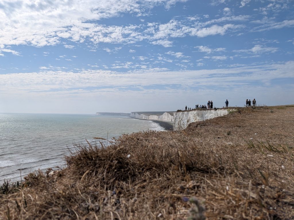 Wide landscape view looking across dry grass and exposed white chalk cliffs toward a group of walkers standing on a distant headland above the sea.
