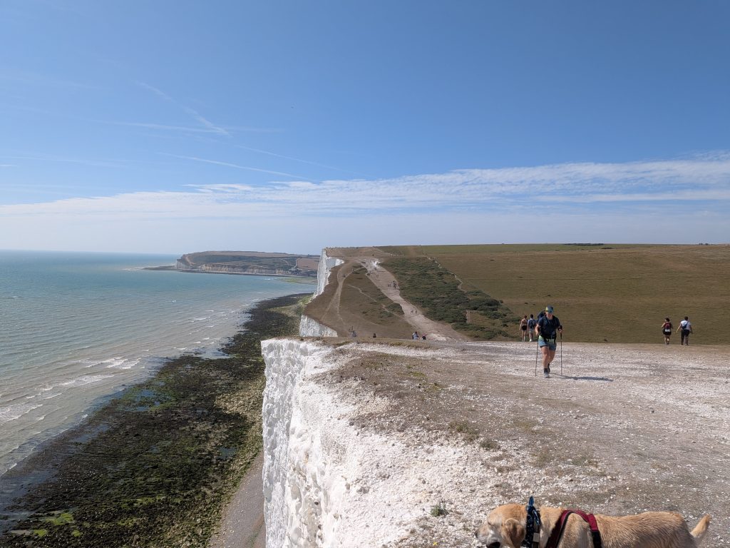 Hiker Syla using trekking poles walking along the cliff-top chalk path at Beachy Head beside trail dog Poppy, above the rocky shoreline and sea.