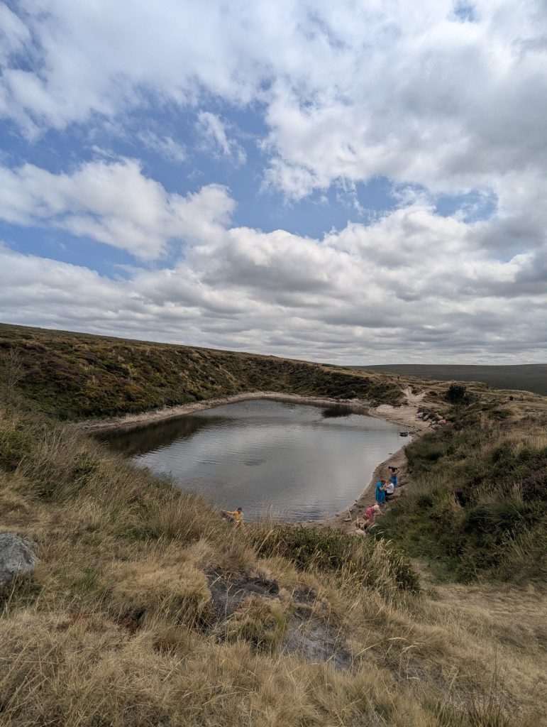 High-angle landscape view of Crazywell Pool, a large flooded tin-mining pool on Dartmoor, surrounded by grassy banks and wild swimmers.