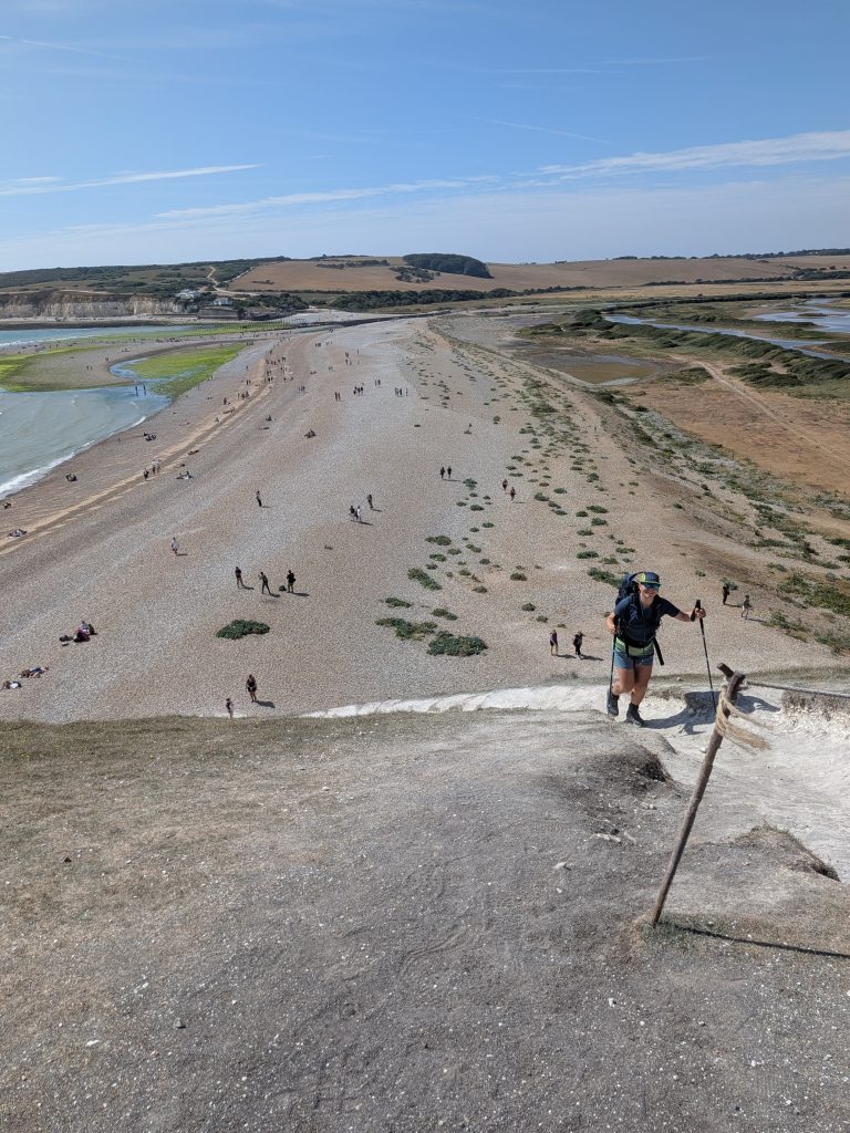 High-angle view looking down a steep chalk path at hiker Syla using trekking poles, with the wide Cuckmere Haven shingle beach and river mouth below.