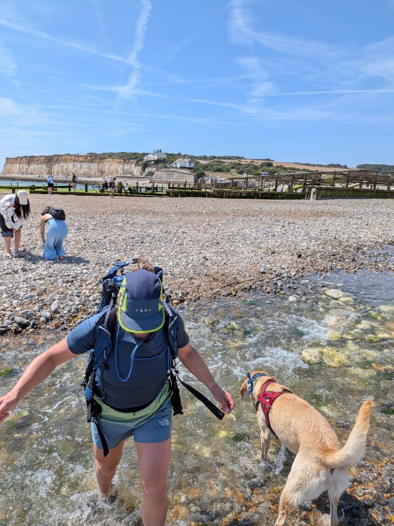 Hiker Syla wearing a VÅGA running cap and rucksack wading through the shallow Cuckmere River mouth at low tide alongside trail dog Poppy.