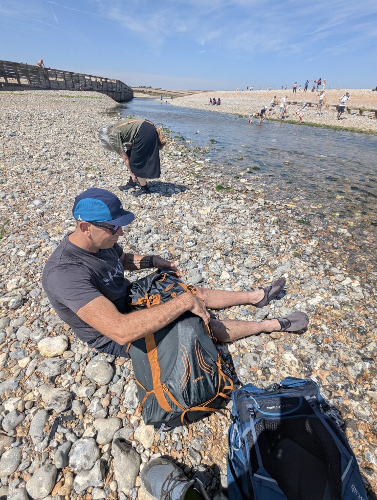 Hiker Terry sitting on the shingle beach at Cuckmere Haven adjusting his Osprey Exos rucksack and water shoes next to the river mouth.