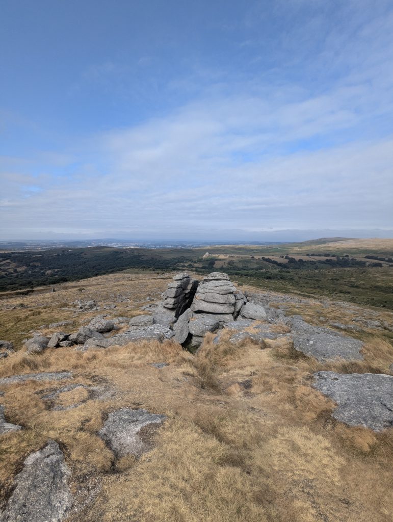 Weathered grey granite boulders stacked naturally on a grassy ridge overlooking rolling moorland under a bright blue sky.