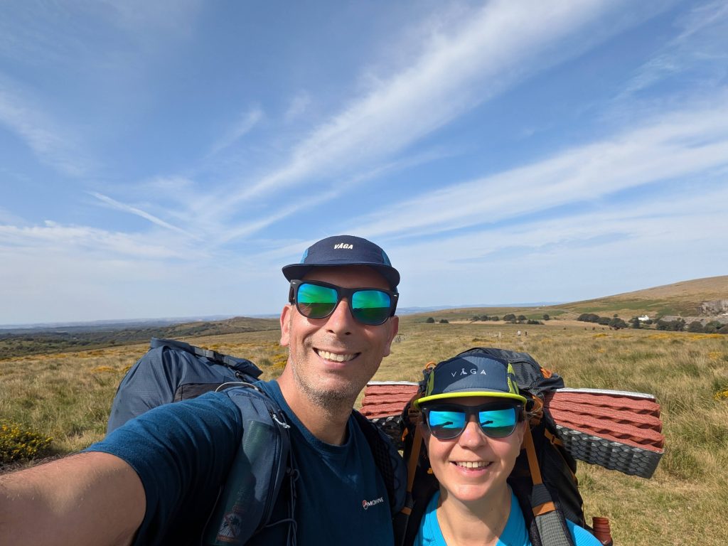 Two smiling hikers wearing VÅGA caps, sunglasses, and technical backpacks under a bright blue sky on Dartmoor.