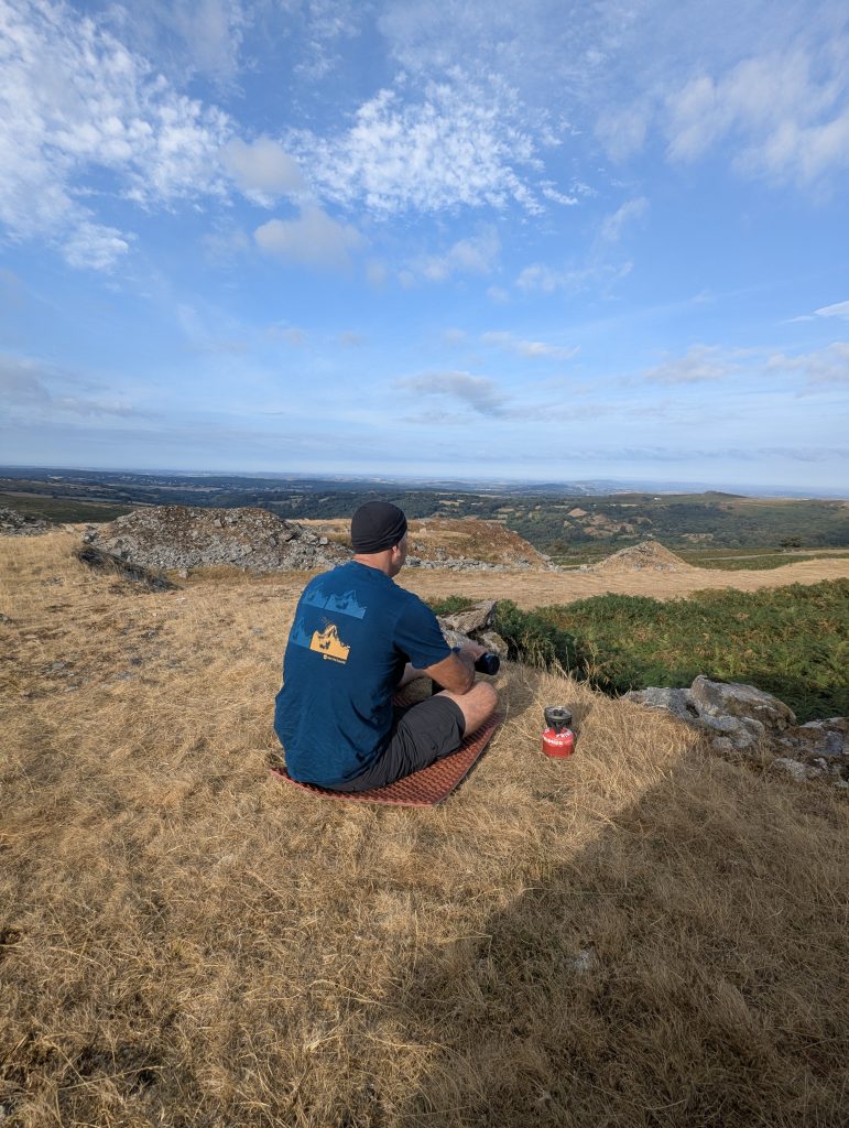 Terry sitting on a red foam sleeping mat beside a Primus trail stove, drinking morning coffee while looking over Swelltor Quarry toward King's Tor on Dartmoor National Park.