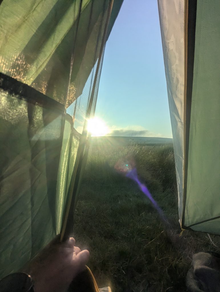 First-person perspective looking out through a green tent door at the bright morning sun rising over dew-covered moorland grass on Dartmoor.