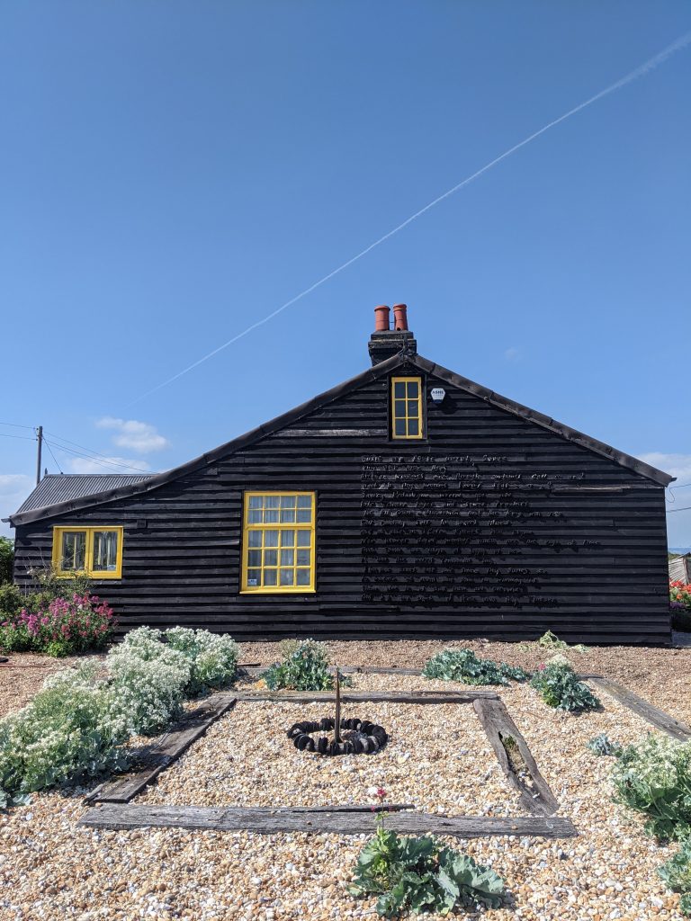 Derek Jarman's Prospect Cottage featuring black timber cladding, bright yellow window frames, and coastal plants.
