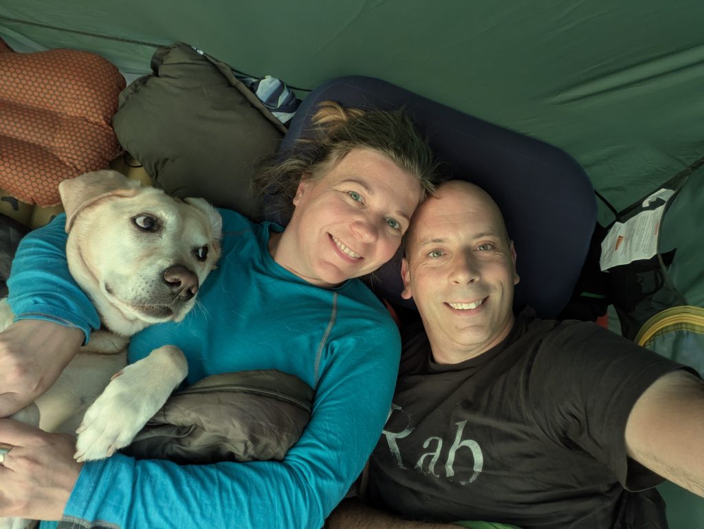 Top-down selfie inside a green tent showing a male and female hiker lying on inflatable sleeping pads next to their yellow Labrador cross, Poppy.