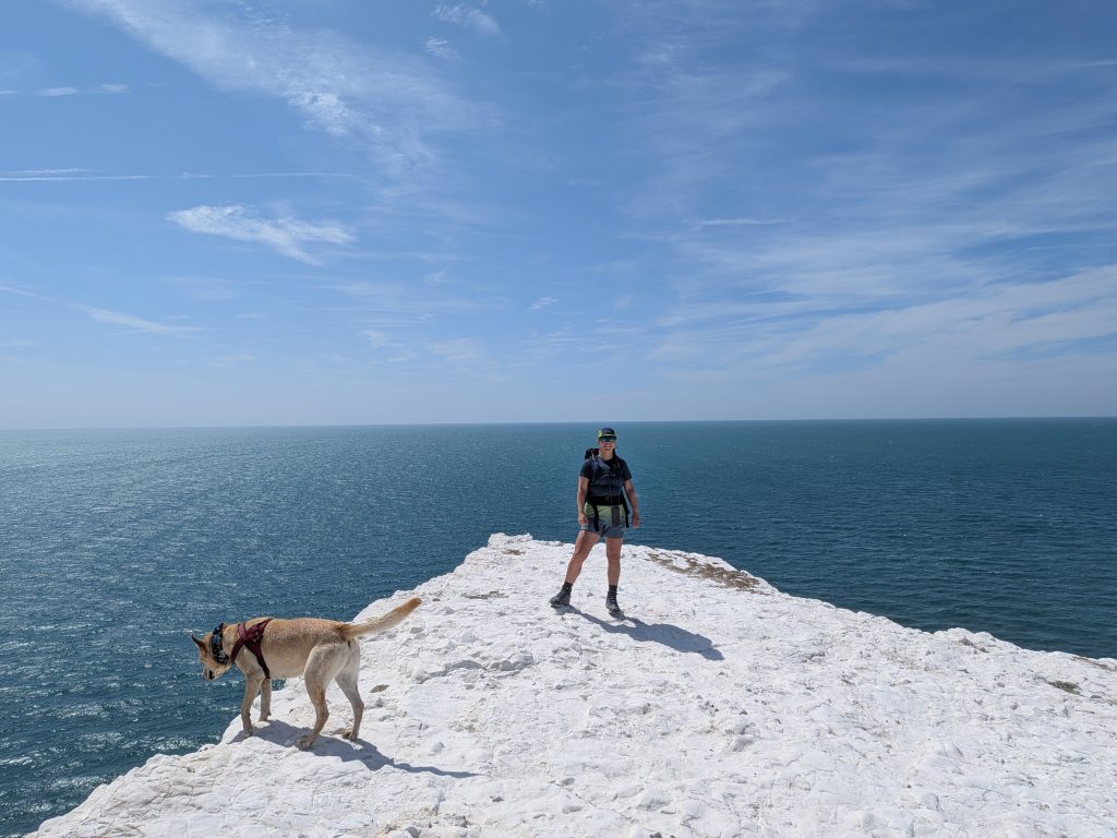 Hiker Syla wearing a VÅGA cap and Rab t-shirt standing on the white chalk summit of Seaford Head beside trail dog Poppy, with the open sea in the background.