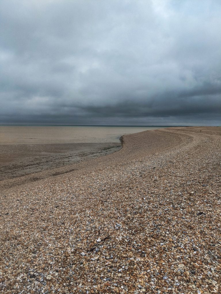 Vast stretch of loose shingle beach meeting the calm ocean water under a moody, overcast sky at Dungeness.