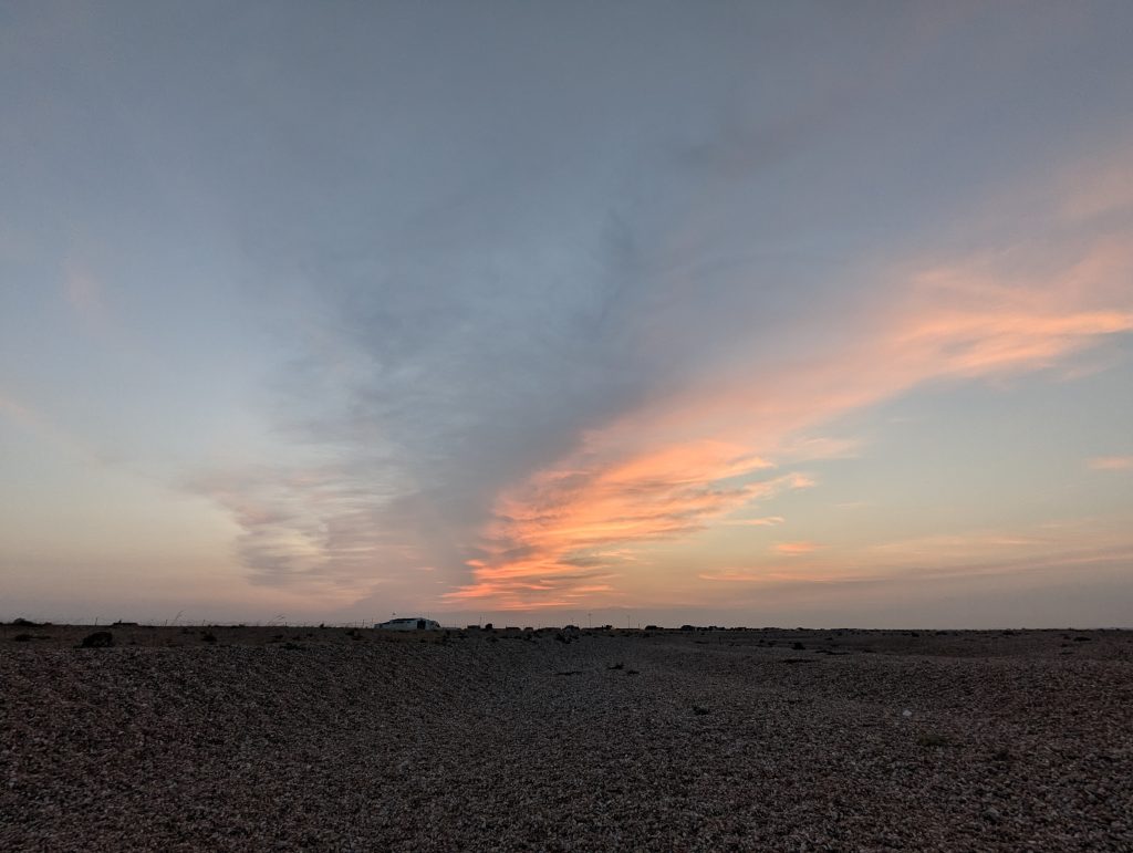 Wide view of the barren Dungeness shingle ridges beneath a dramatic sky painted with sweeping streaks of pink and orange sunset clouds.