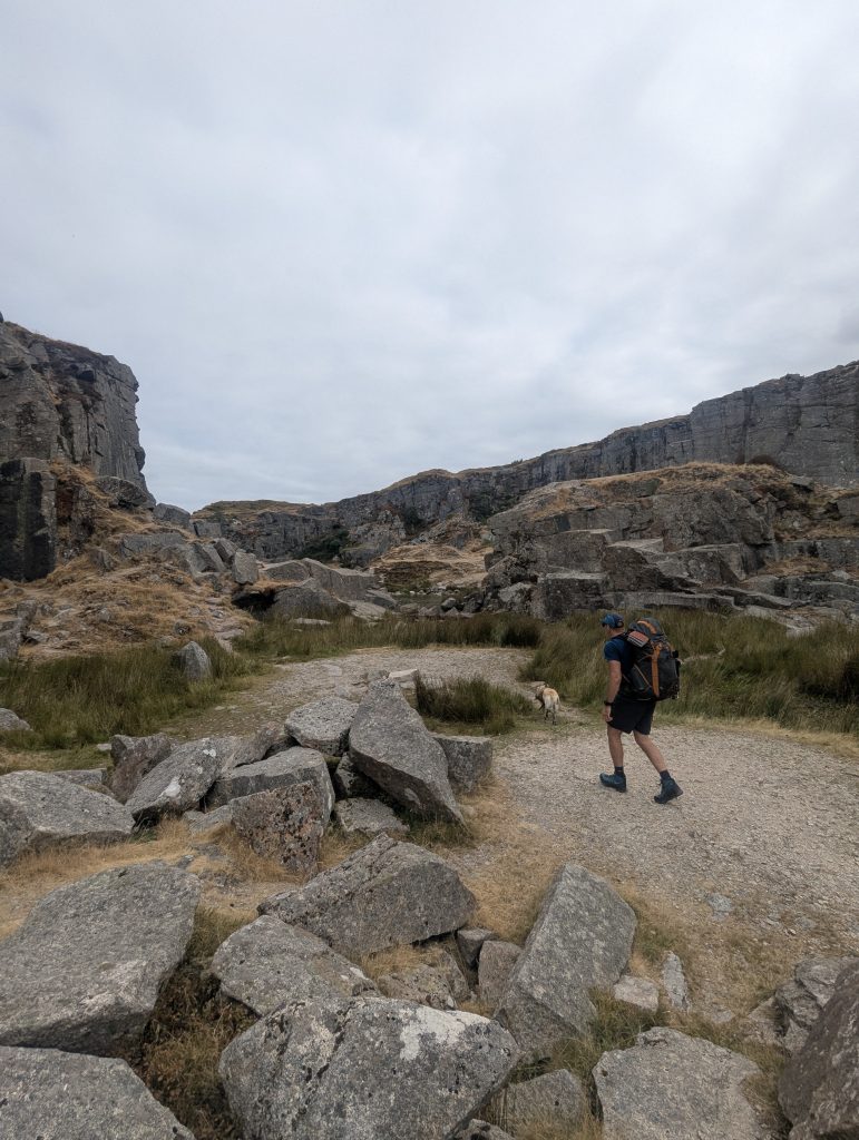 Terry wearing a rucksack walking along a gravel trail into the granite amphitheatre of Foggintor Quarry accompanied by yellow Labrador cross Poppy.