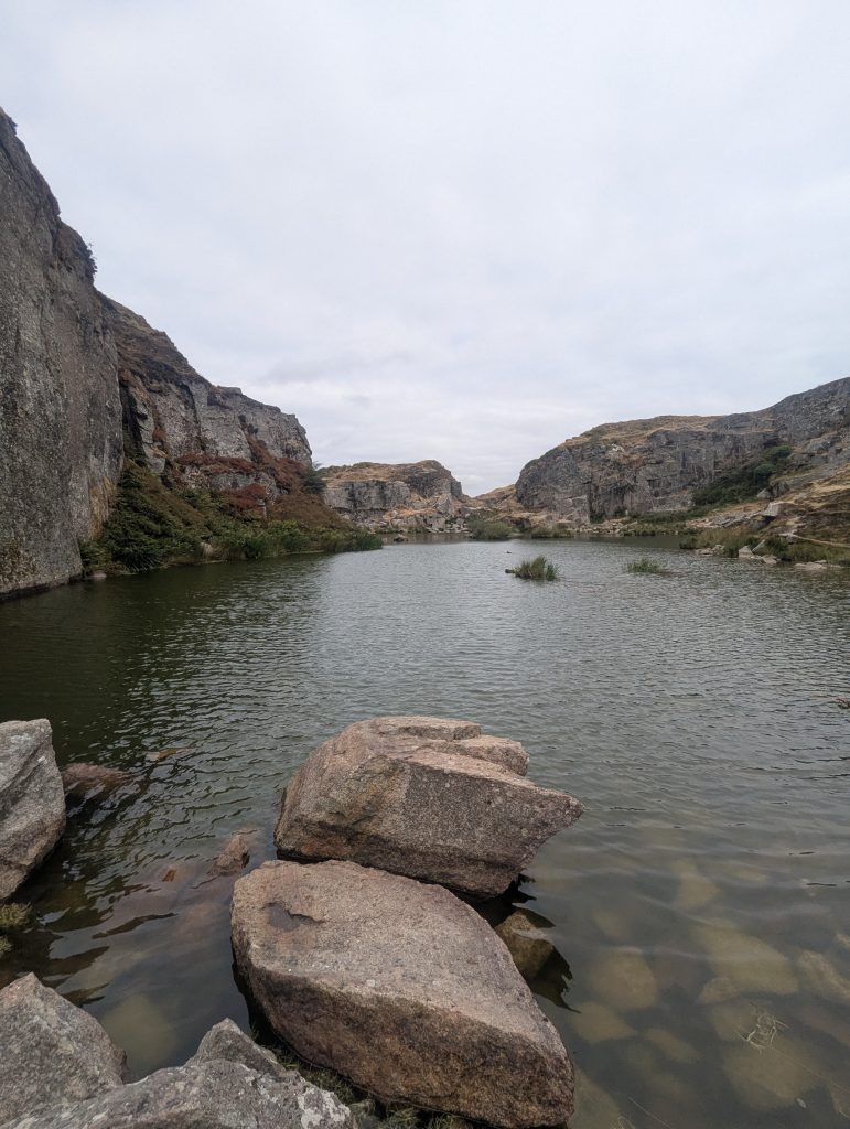 Looking across the calm flooded water basin of Foggintor Quarry surrounded by high granite cut cliffs on Dartmoor under an overcast sky.