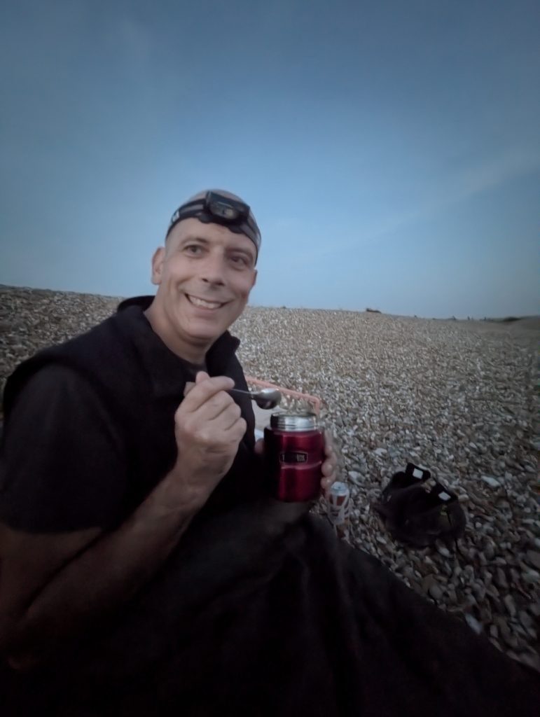 Male hiker wearing a black fleece and headlamp smiling while eating a hot meal from a red insulated food flask on the Dungeness shingle at twilight.