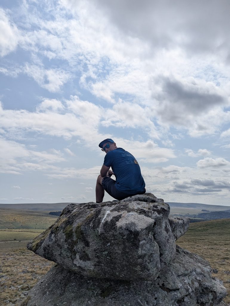 Hiker wearing a blue shirt and cap seated on top of a weathered granite boulder at King's Tor, overlooking Dartmoor National Park.