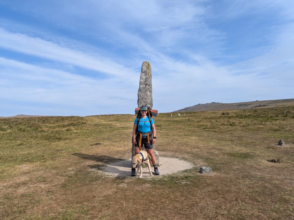 Female backpacker in a blue shirt standing beside the 3-meter Merrivale Menhir standing stone with a yellow Labrador cross.