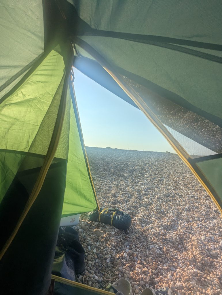 View looking out through the unzipped mesh door of a green camping tent onto a vast expanse of sunlit Dungeness shingle.