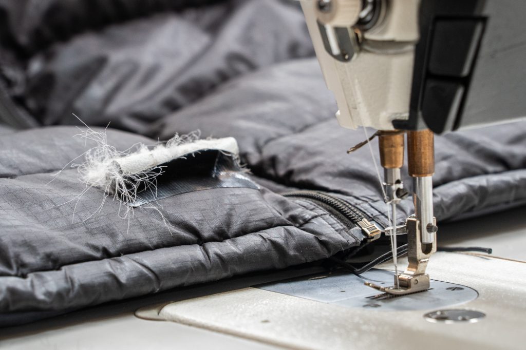 A close-up of an industrial sewing machine needle stitching a repair patch onto a dark grey quilted down jacket, with white insulation spilling from the tear.