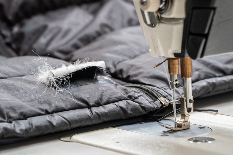 A close-up of an industrial sewing machine needle stitching a repair patch onto a dark grey quilted down jacket, with white insulation spilling from the tear.