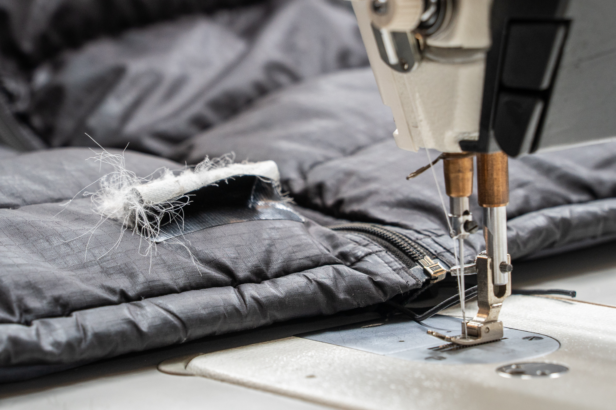 A close-up of an industrial sewing machine needle stitching a repair patch onto a dark grey quilted down jacket, with white insulation spilling from the tear.