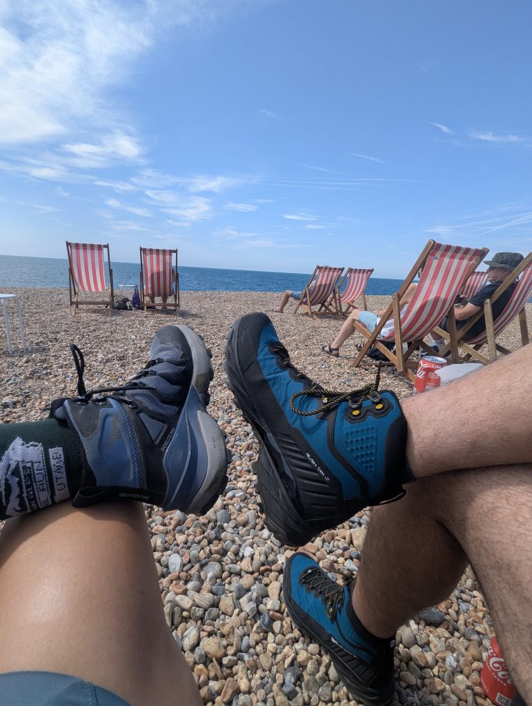Low-angle POV shot showing two hikers relaxing in deck chairs on Seaford shingle beach, displaying inov-8 and Scarpa technical hiking boots against the pebbles and sea.