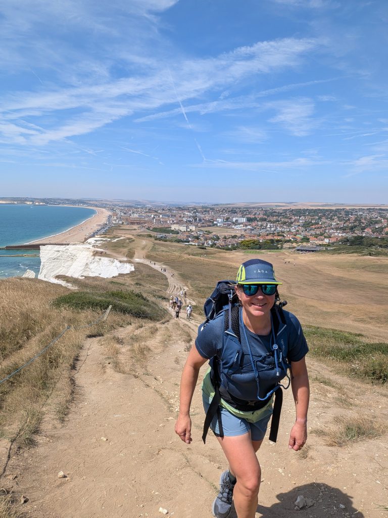Hiker Syla wearing a VÅGA cap and Rab t-shirt climbing a steep, dry dirt trail up Seaford Head, with the town of Seaford and the coastline stretching out below.