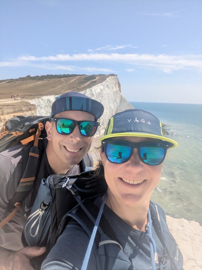 Hikers Syla and Terry smiling for a summit selfie on Seaford Head wearing VÅGA running caps and Osprey rucksacks, with Cuckmere Haven and the Seven Sisters in the background.