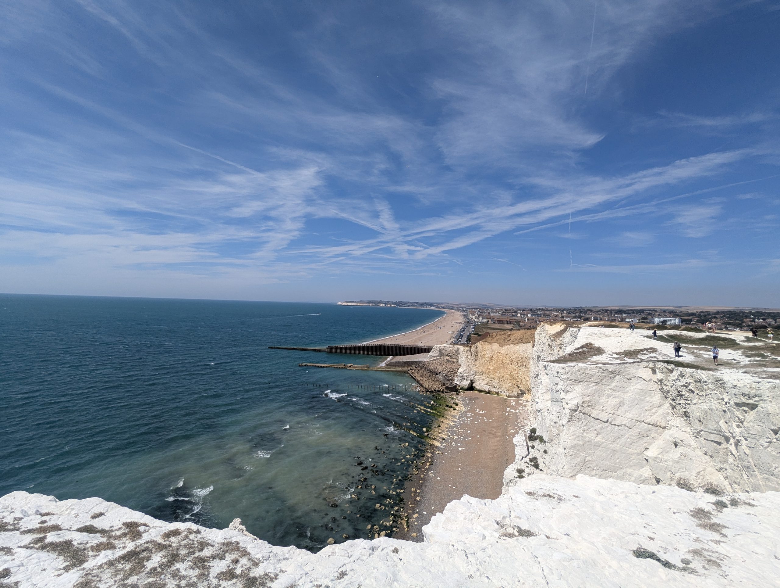 Panoramic high-angle view looking down from the sheer white chalk edge of Seaford Head over the shingle beach and ocean bay of Seaford under a streaky blue sky.