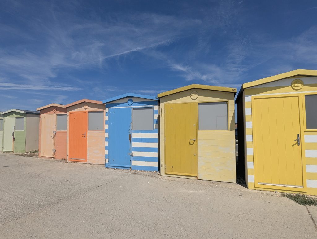 A row of brightly painted colorful beach huts in green, orange, blue, and yellow along the concrete seafront promenade at Seaford.
