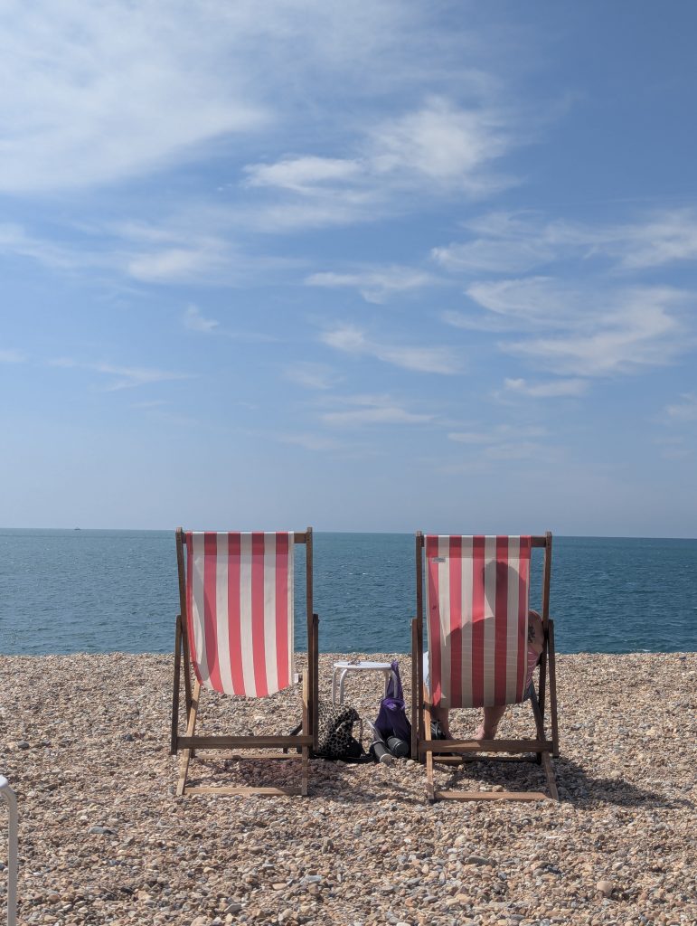 Two red-and-white striped wooden deck chairs set up on the shingle beach at Seaford facing out toward the calm blue sea under a sunny sky.