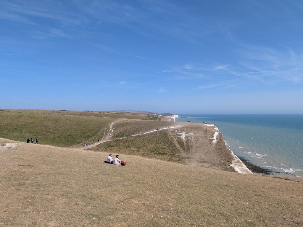 Two hikers sitting on a grassy hilltop slope overlooking the rolling white chalk brows of the Seven Sisters cliffs dropping into the blue sea.
